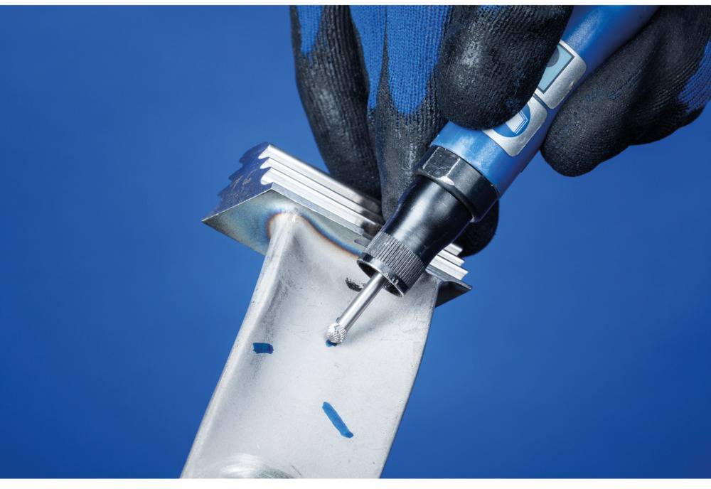 Eine Hand mit Handschuh hält ein silbernes Metallstück und verwendet ein blaues Werkzeug zum Entgraten der Kante. Hintergrund ist blau.