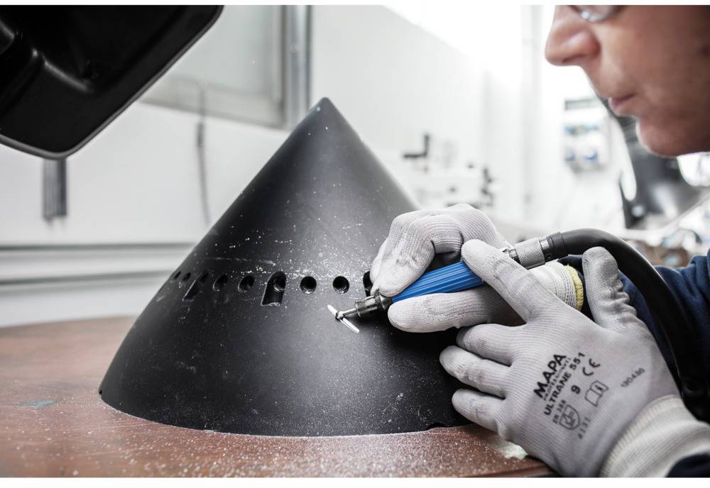 Eine Person bearbeitet mit einem Elektrowerkzeug eine große, schwarze, konische Metallform. Sie trägt Handschuhe und Schutzbrille.