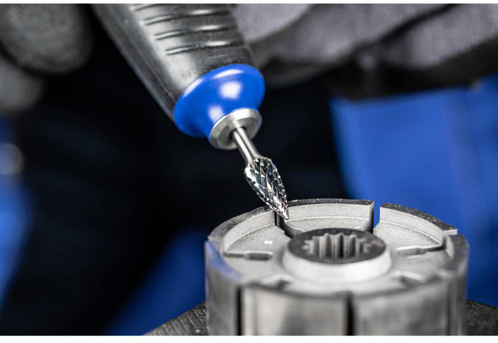 Nahaufnahme eines Drehwerkzeugs mit Schleifkopf, das ein metallisches Zahnrad bearbeitet. Im Hintergrund sind unscharfe Arbeitshandschuhe zu sehen.