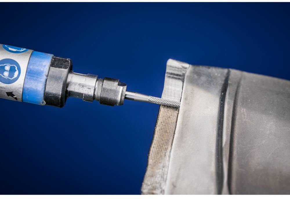 Nahaufnahme eines Bohrers, der auf eine Metallkante gerichtet ist, möglicherweise für Schneide- oder Polierarbeiten, vor blauem Hintergrund.