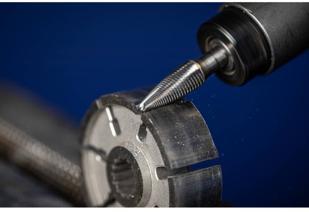 Ein Fräser bearbeitet ein Metallteil mit einem kegelförmigen Bohrer. Das Bild zeigt präzise Metallbearbeitung vor blauem Hintergrund.