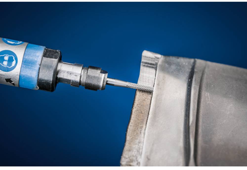 Ein Werkzeug bearbeitet die Kante eines Metallteils. Der Hintergrund ist blau, und das Werkzeug zeigt Symbole für Arbeitssicherheit.