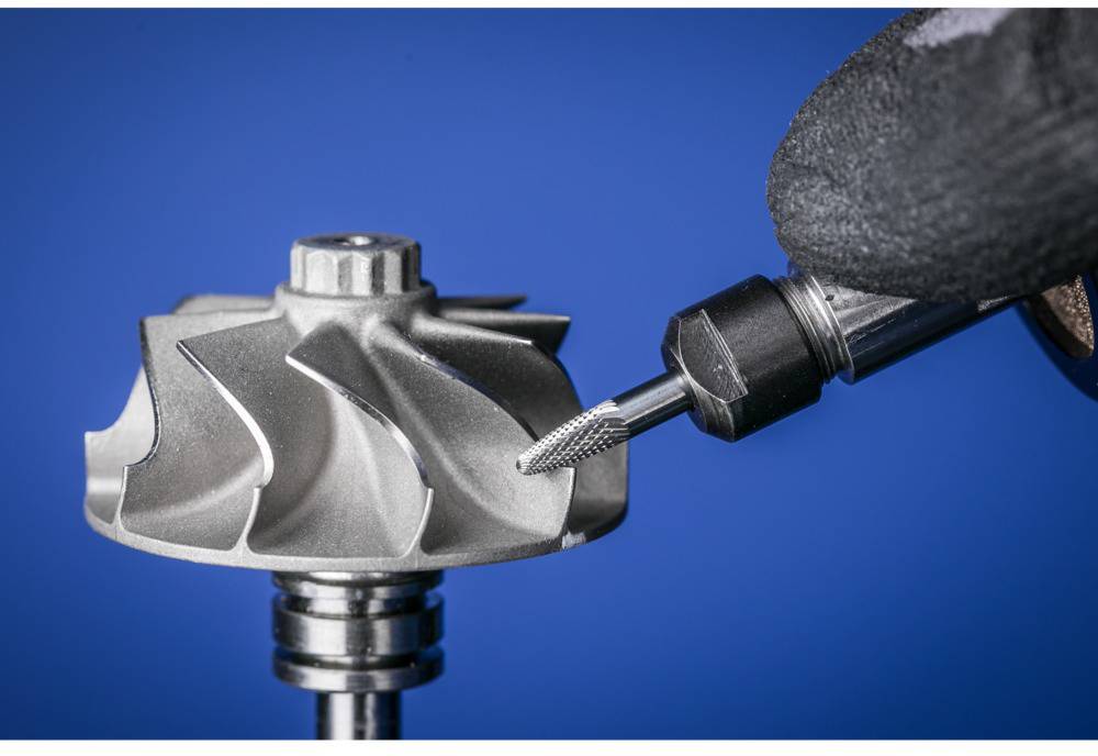 Eine Hand mit Handschuh hält ein Werkzeug und bearbeitet ein metallisches Bauteil mit Schaufeln vor blauem Hintergrund.