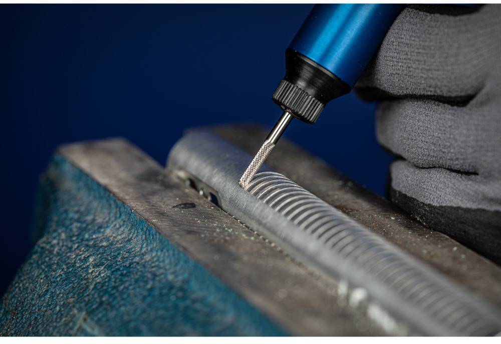 Hand hält ein rotierendes Werkzeug und bearbeitet Metall in einer Schraubzwinge, Detailaufnahme von präziser Glättungsarbeit.