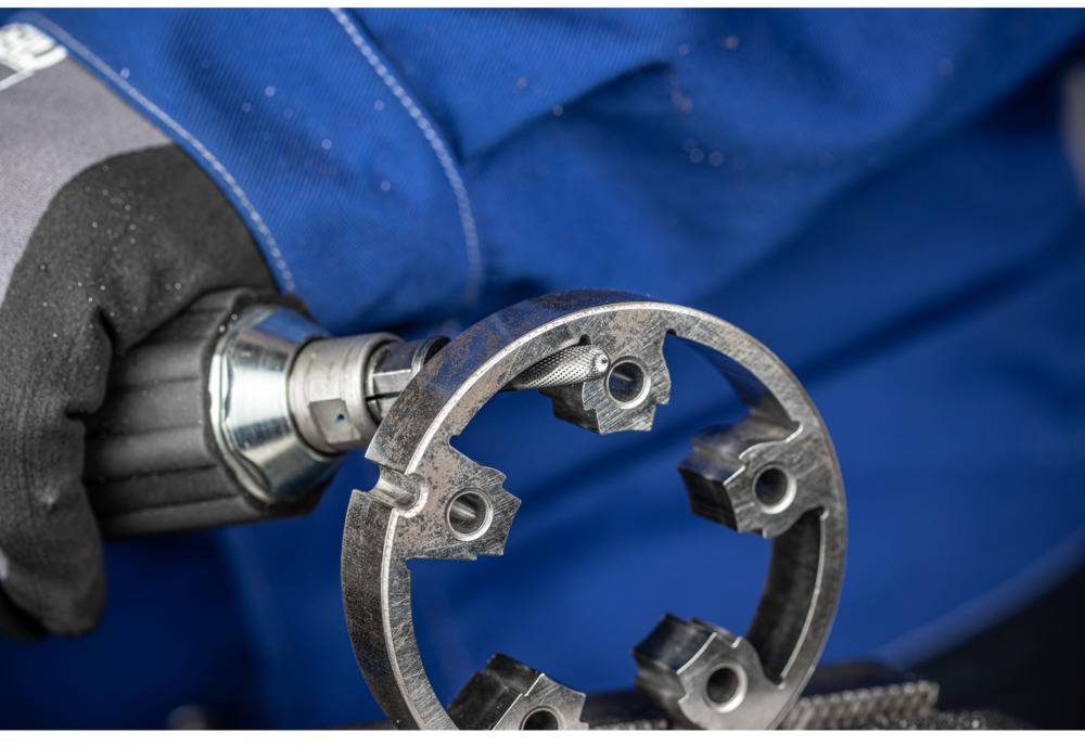 Eine Person in blauer Kleidung verwendet ein Elektrowerkzeug, um Metallbearbeitung an einem runden, mehrspännigen Metallstück durchzuführen.