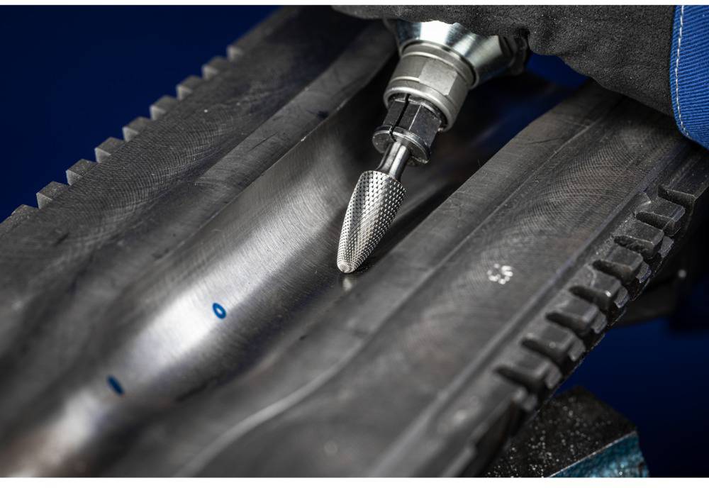 Eine Fräsmaschine bearbeitet eine Metalloberfläche, um Unebenheiten zu entfernen. Eine Hand in einem Handschuh hält das Gerät.