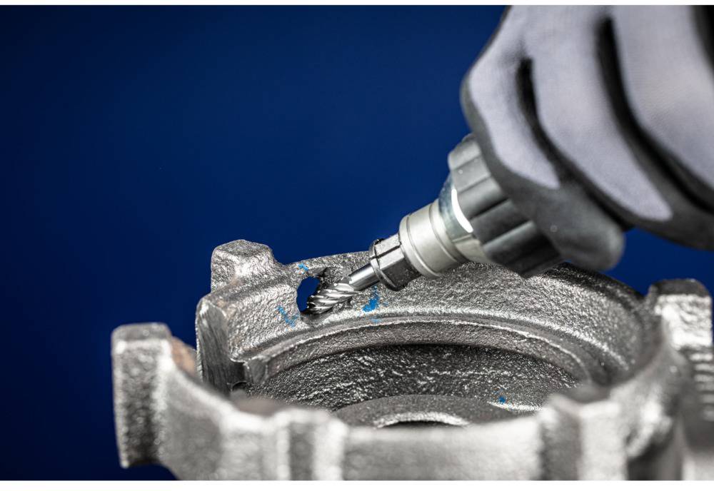 Eine behandschuhte Hand hält ein rotierendes Werkzeug, das Metall von einem Bauteil abschleift. Blauer Hintergrund.