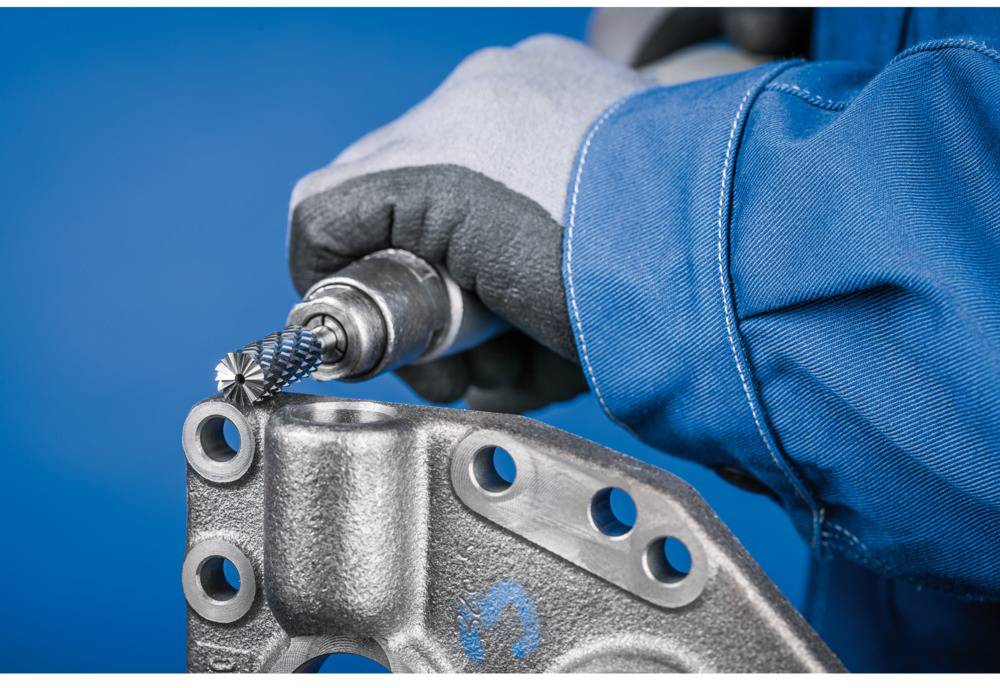 Eine Person trägt Arbeitshandschuhe und benutzt ein Werkzeug, um eine Metallfläche mit Bohrlöchern zu bearbeiten. Das Werkzeug hat einen rotierenden Kopf.