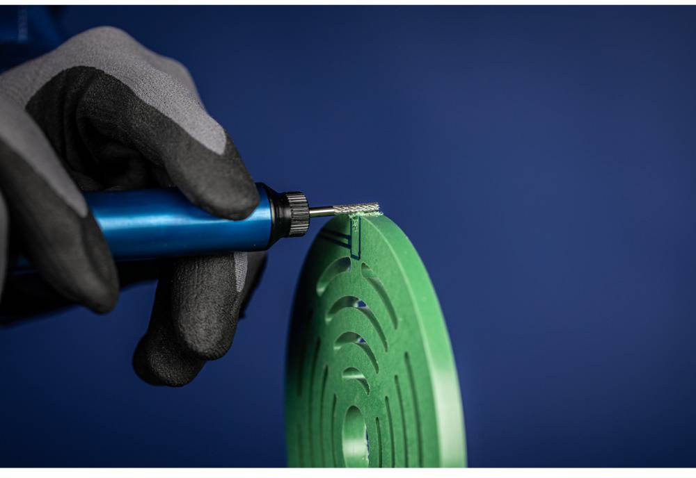 Hand mit Handschuh hält ein rotierendes Werkzeug und bearbeitet ein grünes, kreisförmiges Metallteil vor blauem Hintergrund.