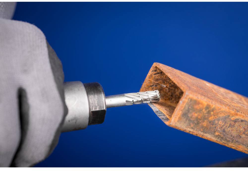 Eine Person, die mit einem Bohrer eine rechteckige, rostige Metallstange bearbeitet. Der Hintergrund ist blau.