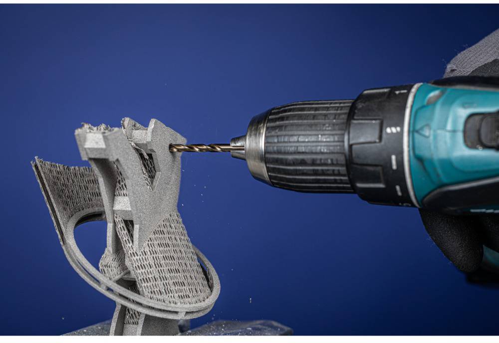 Eine Bohrmaschine bohrt in eine komplexe, dreidimensionale Struktur aus einem grau-schwarzen Material vor blauem Hintergrund.