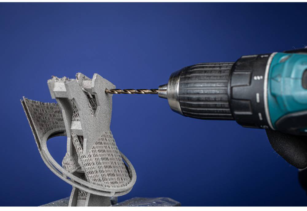 Akkubohrer bohrt in ein komplex geformtes, graues 3D-Druckobjekt vor blauem Hintergrund.