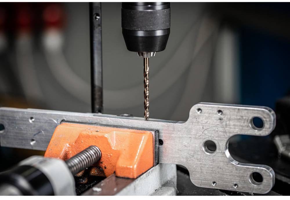 Ein Bohrer durchbohrt ein Metallstück, das in einem Schraubstock eingespannt ist. Das Bild zeigt einen Werkstattvorgang.