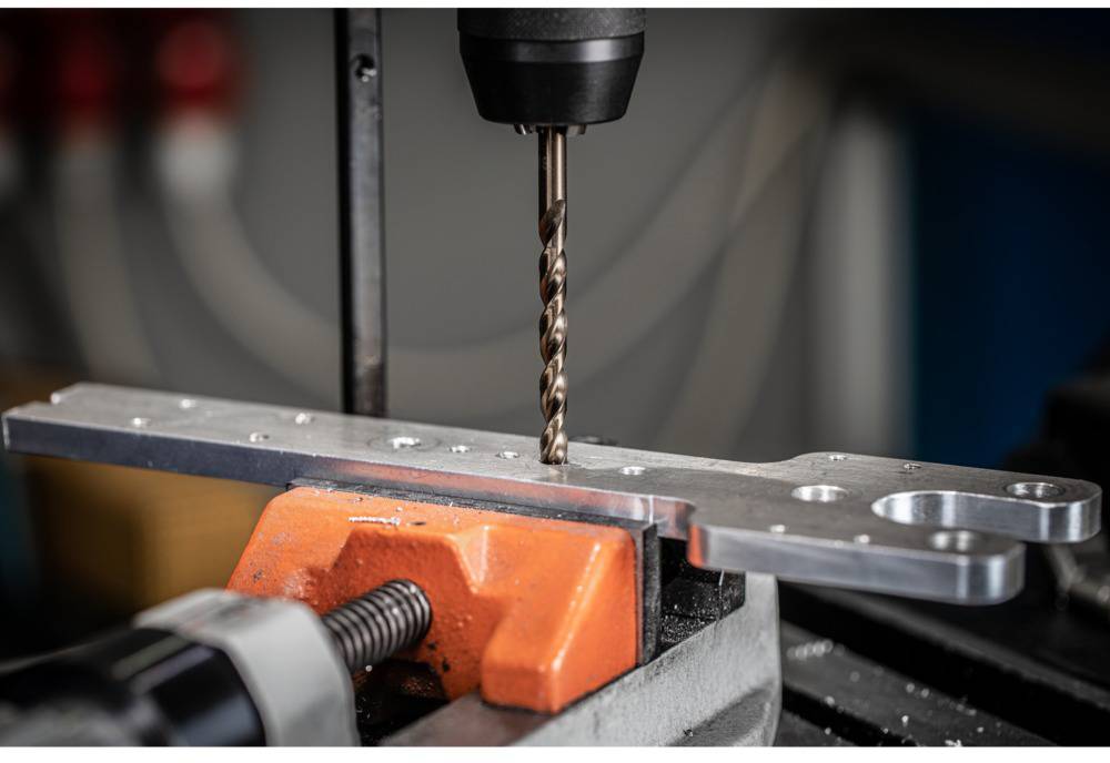 Ein Bohrer bohrt Löcher in eine Metallplatte, die in einem Schraubstock gehalten wird. Der Hintergrund zeigt eine Werkstattumgebung.