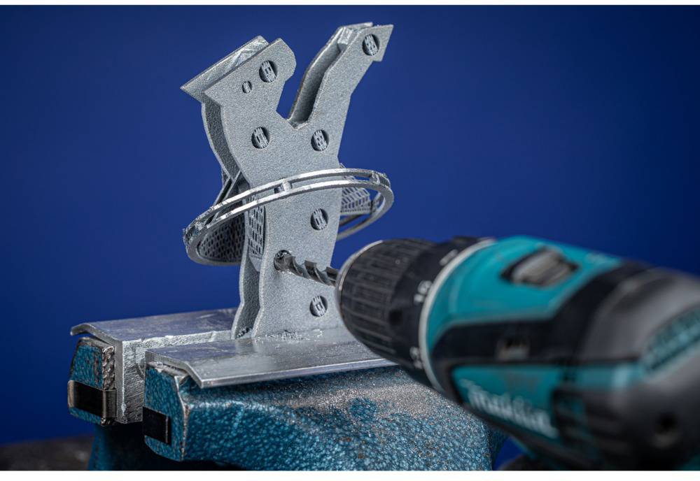 Eine Bohrmaschine bearbeitet ein metallisches Werkstück, das in einem Schraubstock befestigt ist. Der Hintergrund ist blau.