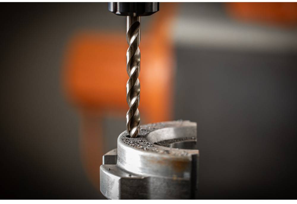 Nahaufnahme eines Bohrers, der ein kreisförmiges Metallstück bearbeitet. Hintergrund unscharf mit orangefarbenem Detail.