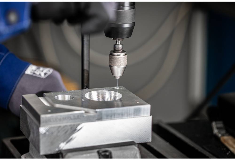 Eine Person bohrt ein Loch in ein Metallstück mit einer Bohrmaschine, während sie spezielle Arbeitshandschuhe trägt.