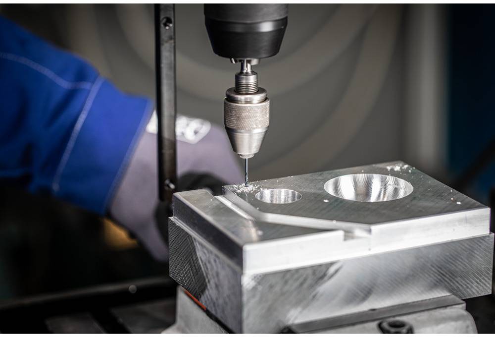 Eine Maschine bohrt präzise Löcher in einen Metallblock. Ein Arbeiter in blauer Schutzkleidung hält das Werkstück stabil.