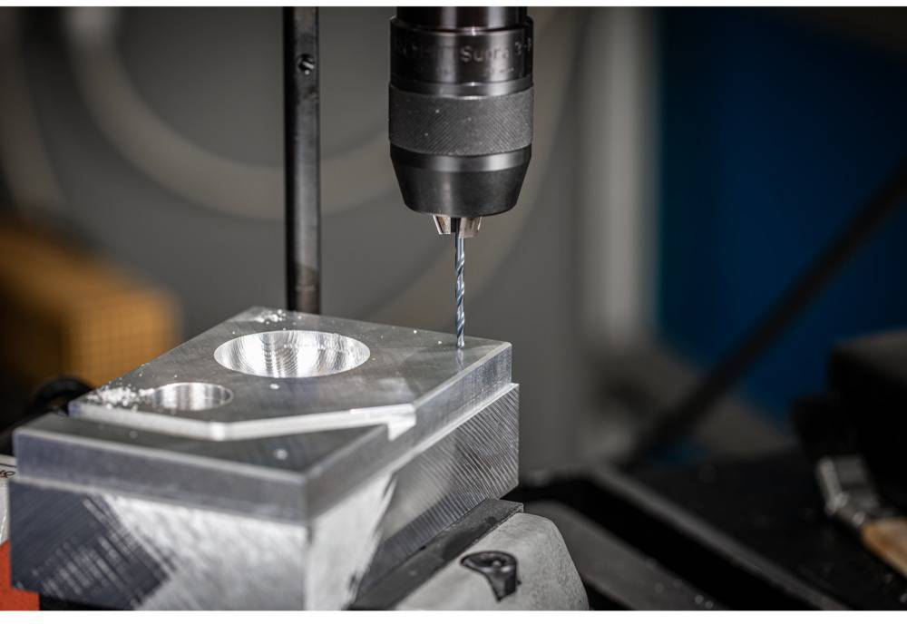 Metallbohrer bearbeitet rechteckigen Metallblock mit Löchern. Nahaufnahme in Werkstattumgebung.