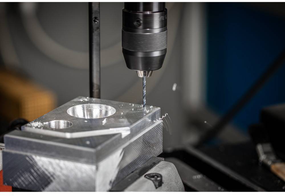 Eine Bohrmaschine bearbeitet ein rechteckiges Metallblockstück mit kreisförmigen Vertiefungen, umgeben von Metallspänen.