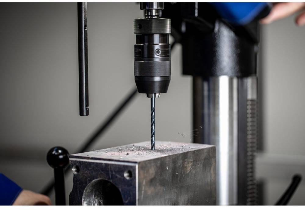 Eine Bohrmaschine bearbeitet ein Metallstück in einer Werkstattumgebung. Metallspäne sind sichtbar um das Bohrloch.