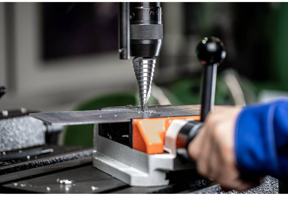 Eine Person benutzt eine Ständerbohrmaschine, um ein Metallblech zu durchbohren. Der Fokus liegt auf dem Bohrvorgang.