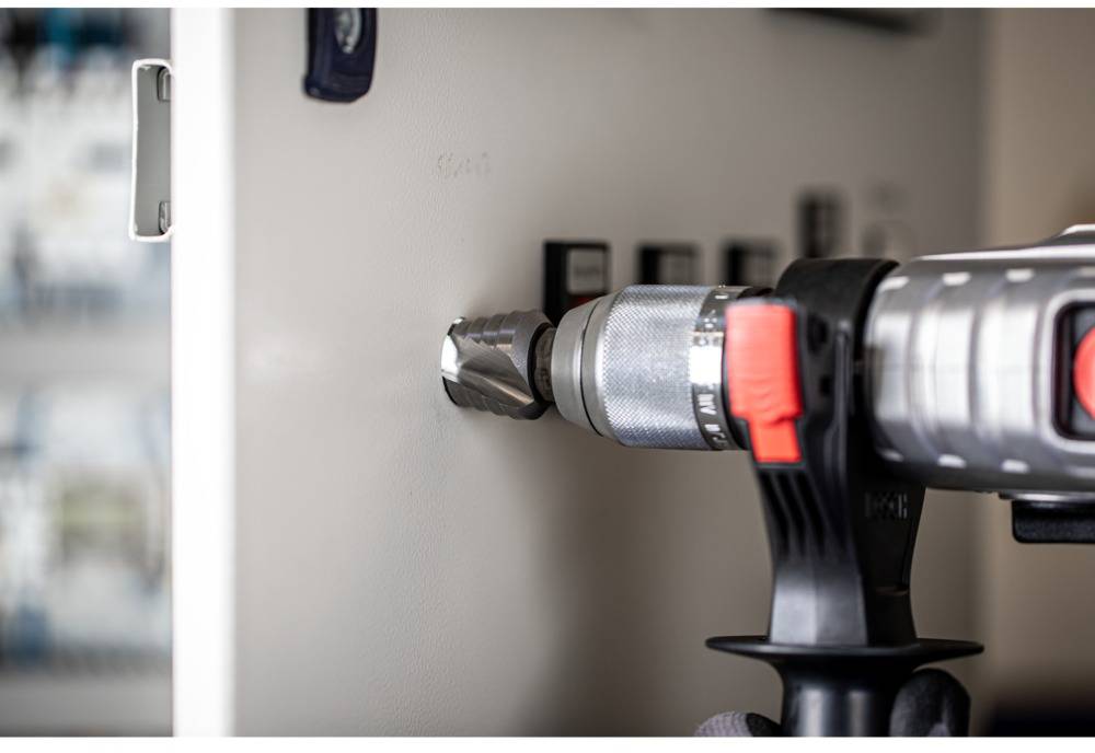 Eine Bohrmaschine durchbohrt die Wand eines elektrischen Schaltkastens. Der Bohrer ist schräg von der Seite sichtbar.