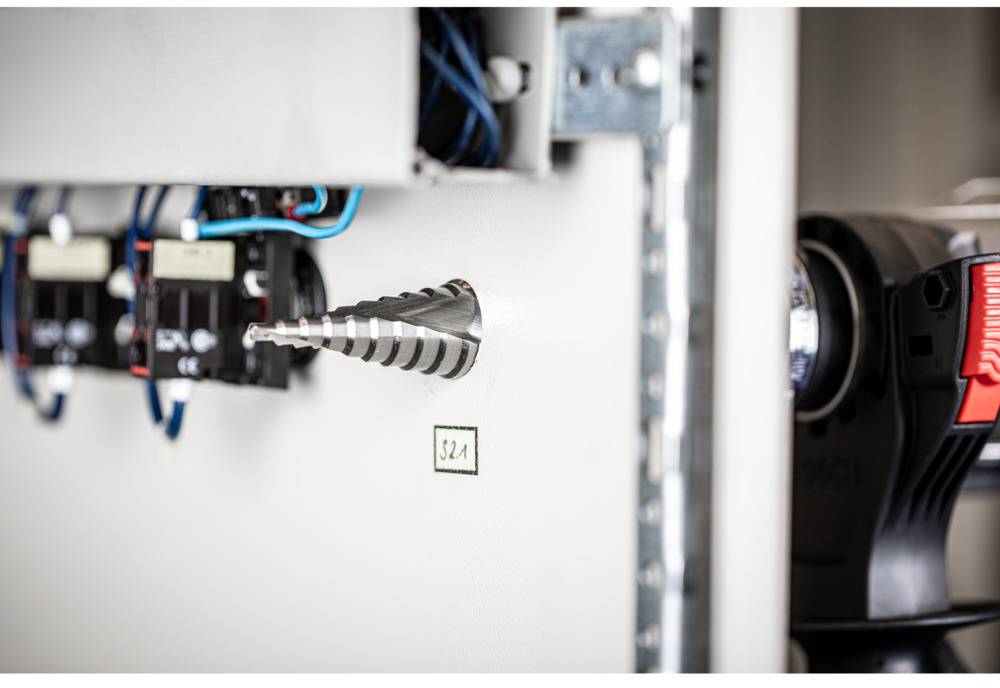 Ein Metallbohrer ist in ein elektrisches Schaltschrankpanel eingeschraubt, um ein Loch zu bohren. Kabel und Schalter sind sichtbar.