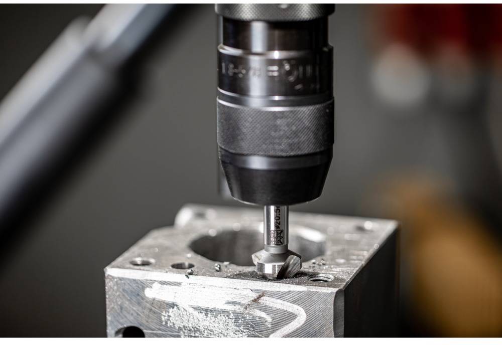 Ein Bohrer bearbeitet ein Metallstück in einer Werkstatt. Im Hintergrund sind unscharfe Werkzeuge zu sehen.