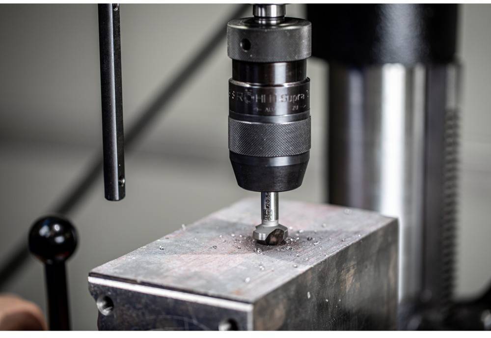 Bohren eines Metallblocks mit einer Ständerbohrmaschine; Metallspäne sichtbar. Arbeitsprozess in einer Werkstatt oder Fertigungsumgebung.