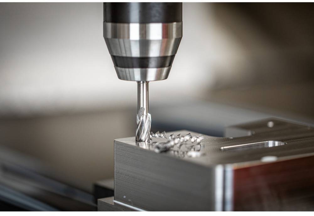 Bohrmaschine bearbeitet Metallblock, erzeugt feine Metallspäne; Nahaufnahme zeigt Präzisionsbearbeitung in Industrieumgebung.
