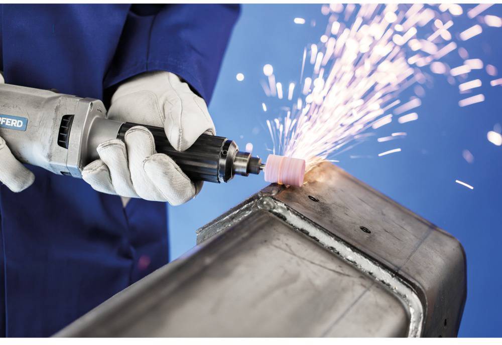 Eine Person schleift mit einem Winkelschleifer Metall, Funken fliegen. Arbeiter trägt blaue Schutzkleidung und weiße Handschuhe.