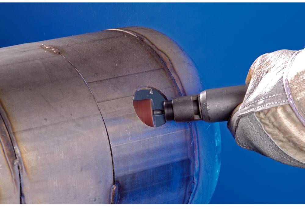 Hand hält Bohrwerkzeug, das ein Loch in ein Metallrohr bohrt. Das Rohr ist zylindrisch und blau beleuchtet.