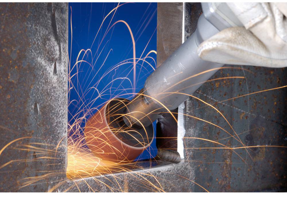 Ein Metallarbeiter schleift mit einem Werkzeug an einer Metallschicht, Funken fliegen in einer geschützten Werkstattumgebung.