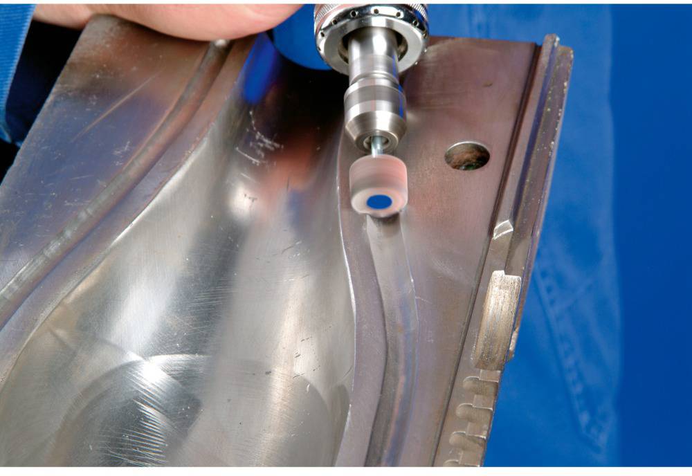 Eine Person poliert die Oberfläche eines Metallteils mit einem rotierenden Werkzeug. Das Metall hat eine gekrümmte Form.