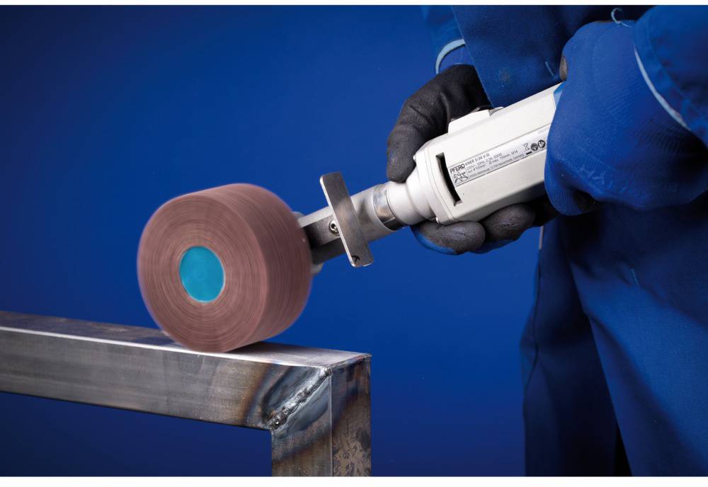 Person schleift ein metallisches Werkstück mit einer elektrischen Schleifmaschine. Hintergrund in Blau gehalten.
