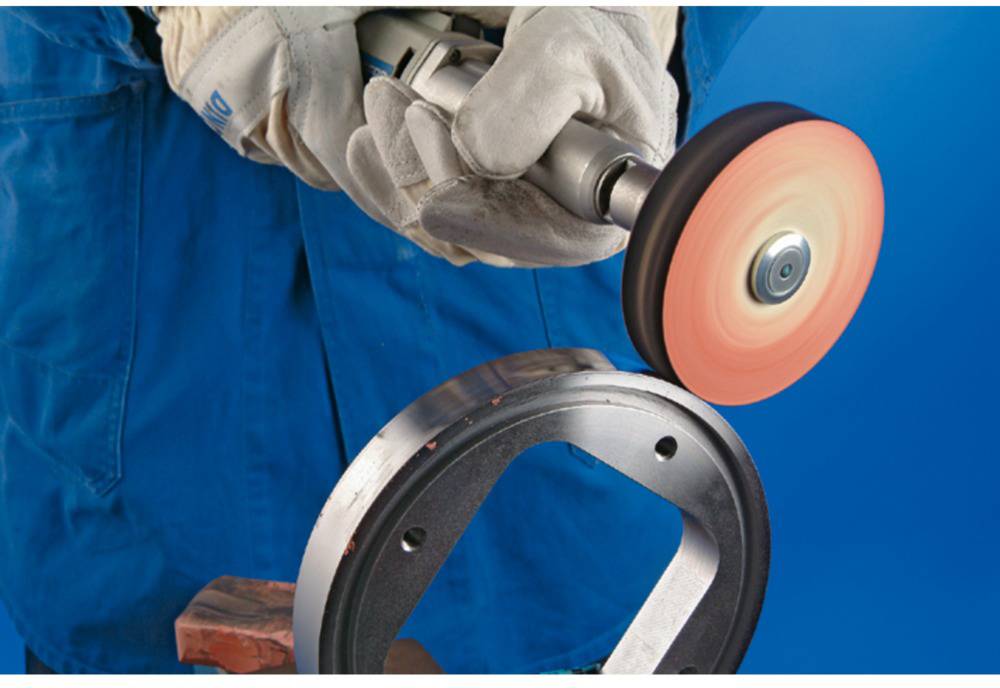 Person schleift ein Metallteil mit einer rotierenden Schleifscheibe, trägt blaue Schutzkleidung und Handschuhe.