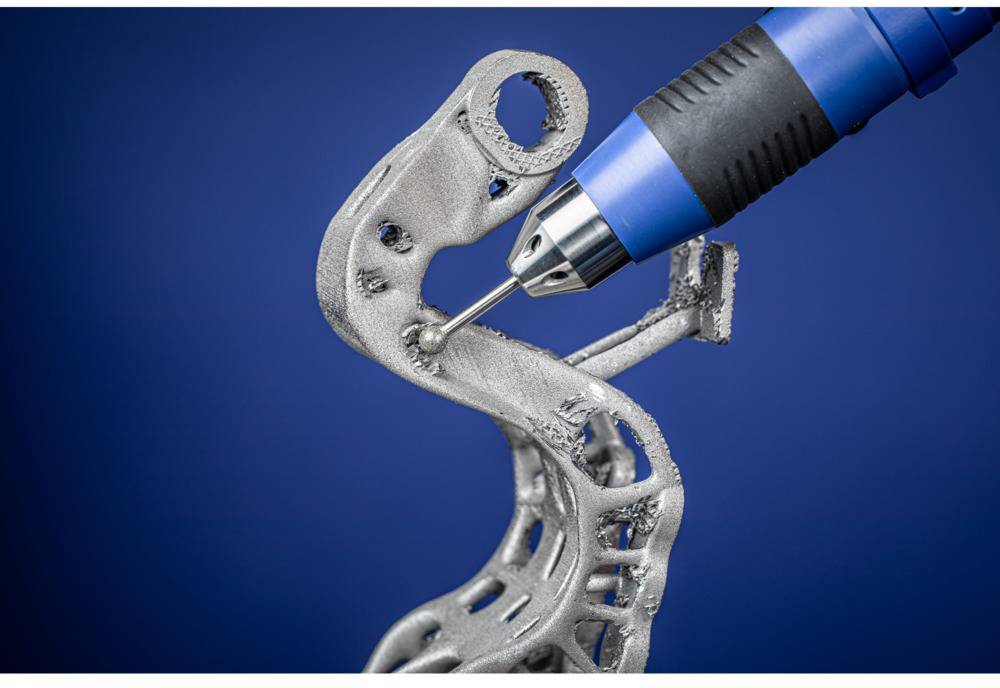 Ein 3D-Drucker mit blauer Düse bearbeitet ein komplexes, metallisches Bauteil vor blauem Hintergrund.