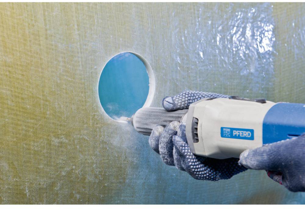 Eine Person trägt Arbeitshandschuhe und bohrt mit einem blauen Werkzeug ein rundes Loch in eine glatte, beige Oberfläche.