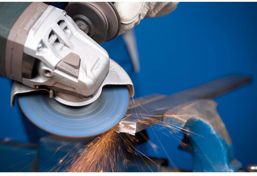 Eine Hand hält einen Winkelschleifer, der Funken von einem Metallstück sprüht, vor einem blauen Hintergrund.