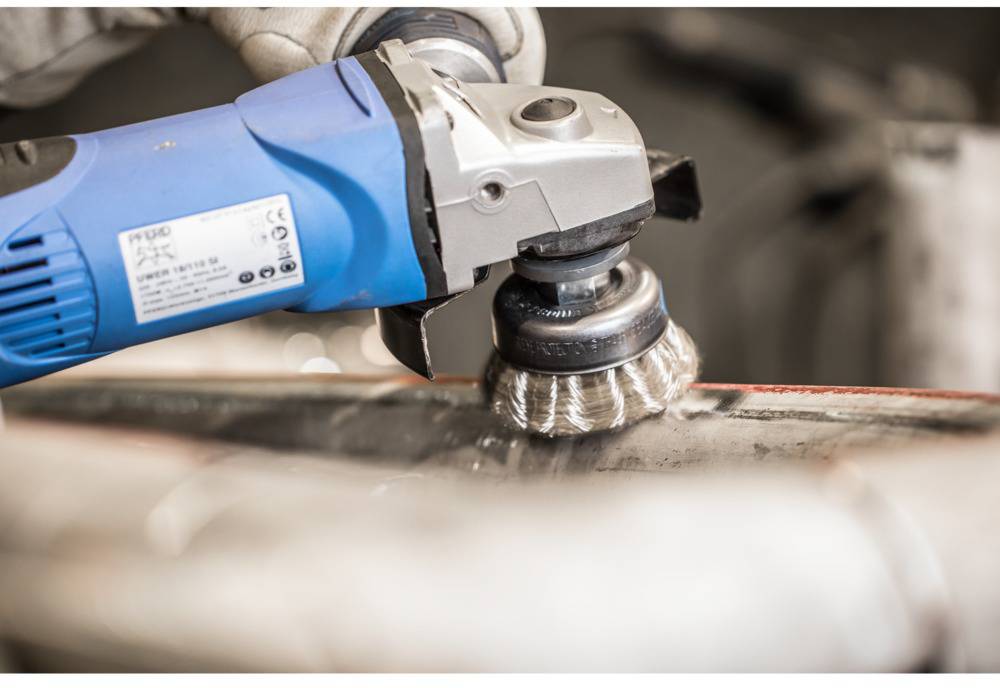 Eine blaue Schleifmaschine mit Drahtbürstenaufsatz schleift Metalloberfläche ab. Die Maschine wird von einer Person in Handschuhen gehalten.