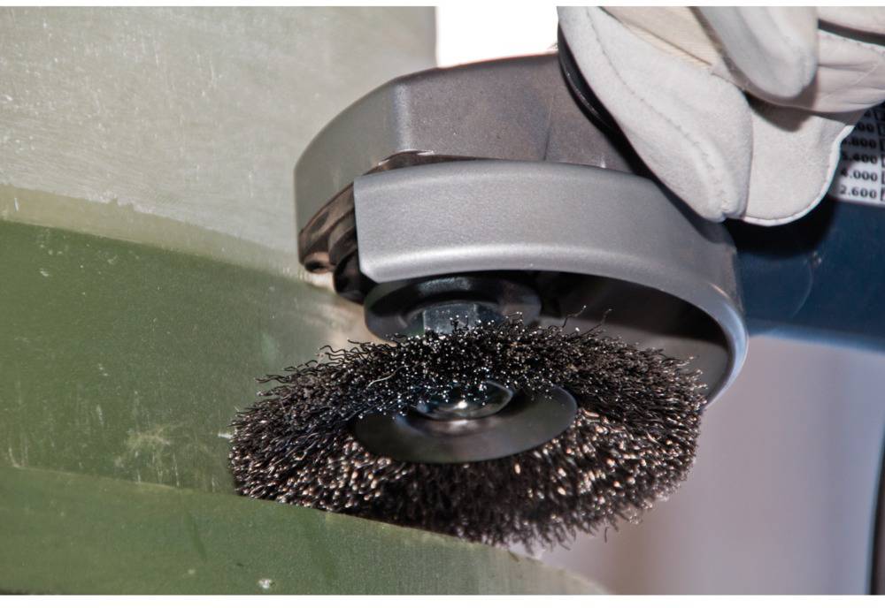 Eine Winkelschleifmaschine mit Drahtbürstenaufsatz bearbeitet eine Oberfläche. Ein Handschuh hält das Werkzeug.