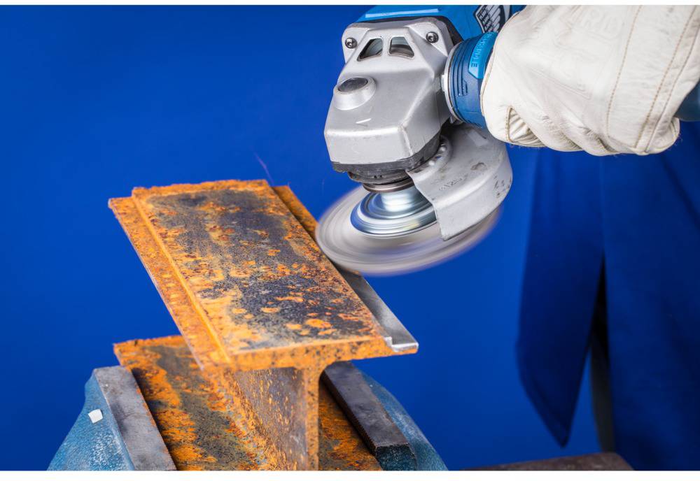Ein Arbeiter schleift mit einer Winkelschleifmaschine einen rostigen Metallbalken. Der Balken wird in einem Schraubstock gehalten.