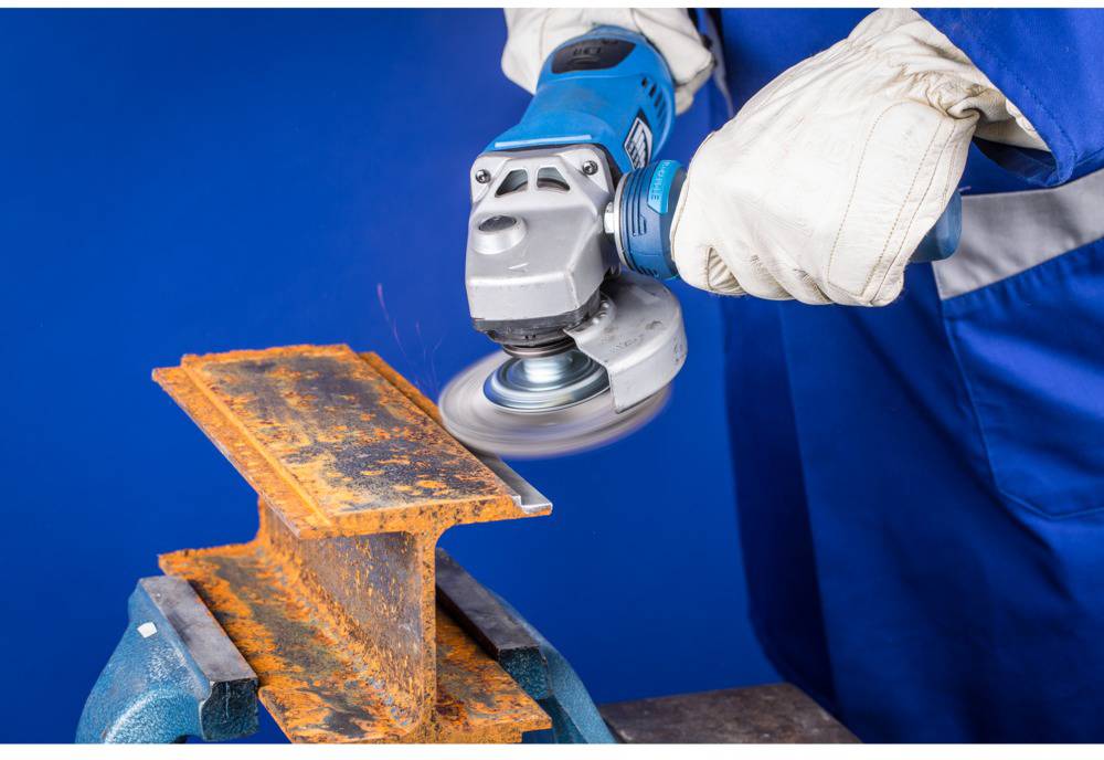 Person mit blauer Arbeitskleidung und Schutzhandschuhen schleift mit einer Winkelschleifer eine verrostete Metallplatte.