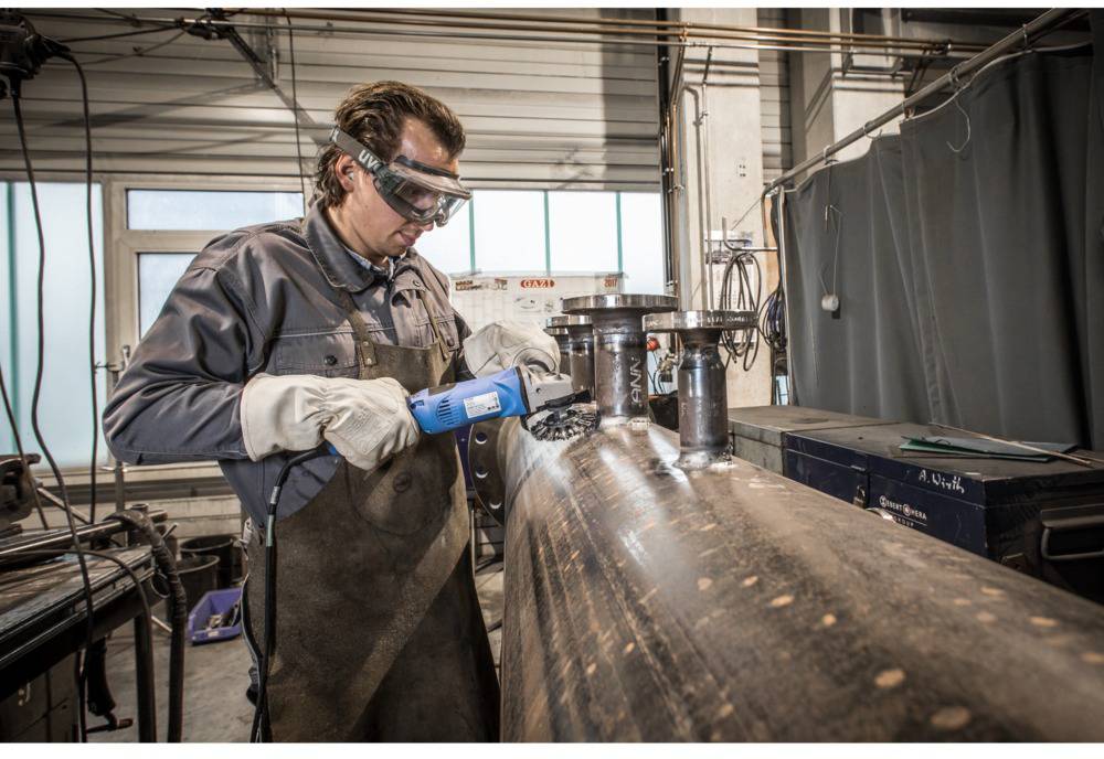 Ein Mann in Schutzkleidung bearbeitet ein großes Metallrohr mit einem elektrischen Werkzeug in einer Werkstattumgebung.