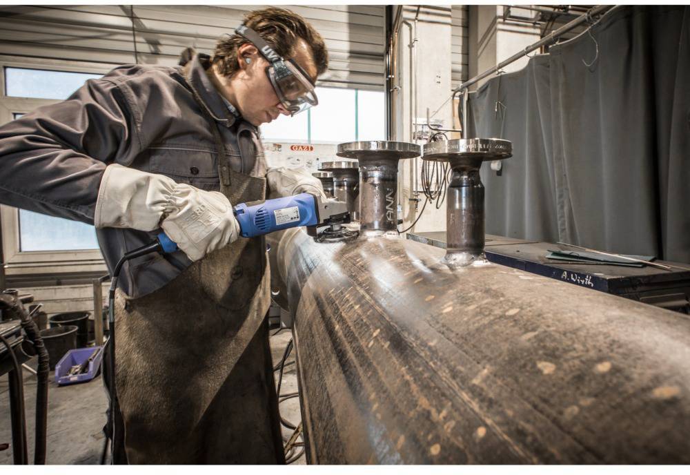 Ein Arbeiter trägt Schutzkleidung und schleift eine Metalloberfläche mit einem Elektrowerkzeug in einer Werkstattumgebung.