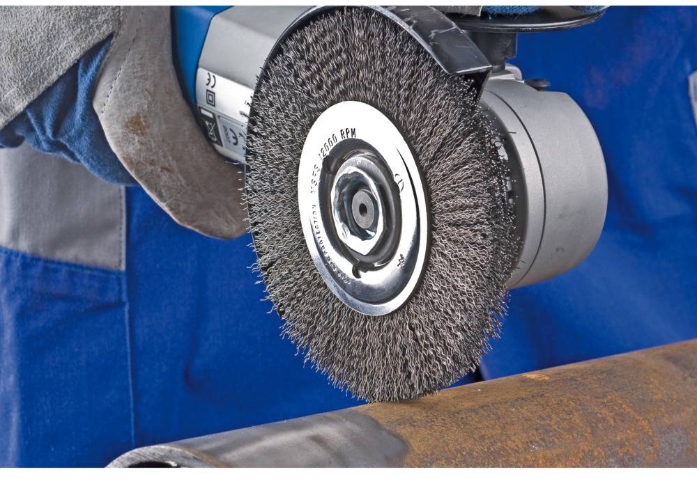 Eine Person schleift mit einer elektrischen Drahtbürste Rost von einem Metallrohr. Die Hand trägt einen Arbeitshandschuh.