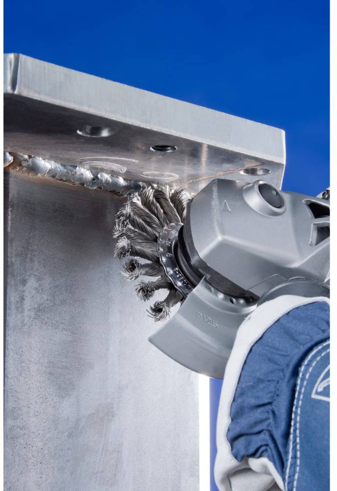 Eine Person mit Schutzkleidung reinigt eine Metallschweißnaht mit einem Schleifgerät vor einem klaren, blauen Hintergrund.