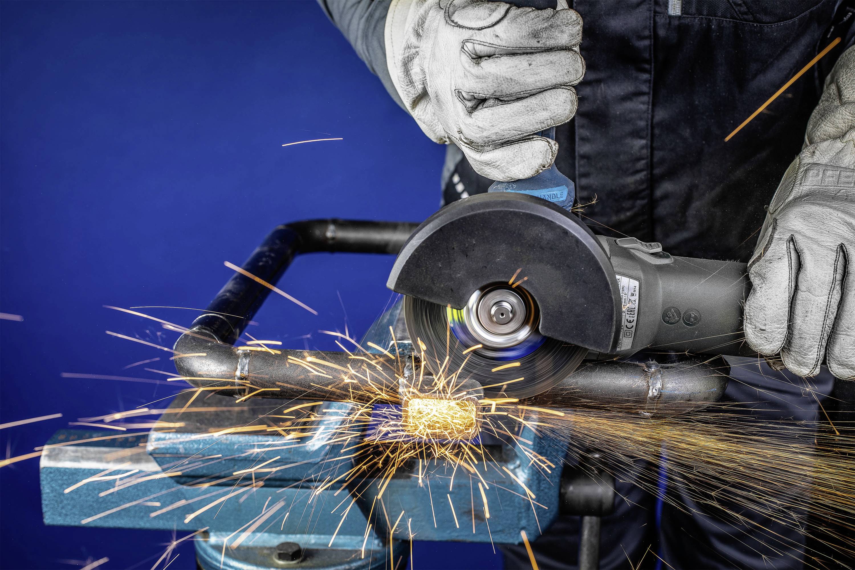 Eine Person trägt Schutzhandschuhe und verwendet einen Winkelschleifer, um ein Metallrohr zu schneiden. Funken sprühen in alle Richtungen.