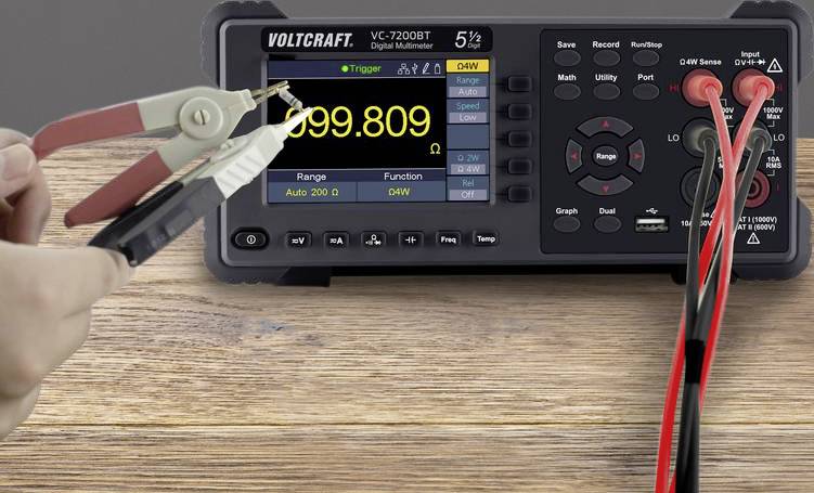 Digitalmultimeter auf Holztisch zeigt '999.809' an. Clip-Messleitungen an Anschlüssen. Links eine Hand beim Messen.